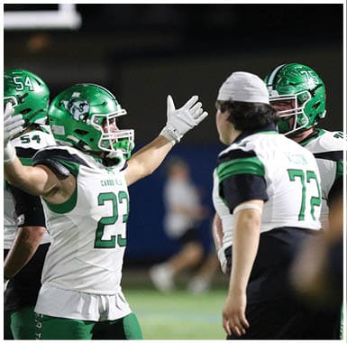 Three people in green football uniforms and helmets stand on a field at night with one person facing away from the camera and the others partially turned. Descripci n generada con IA