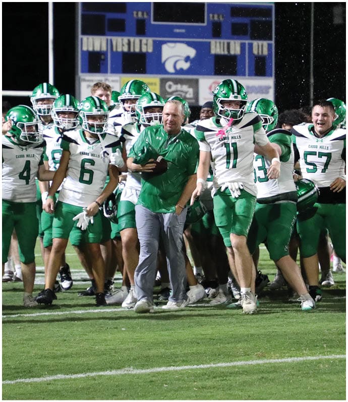 A group of football players in green uniforms are gathered on the field near the end zone while a referee stands in the foreground and the scoreboard shows the home team leading. Descripci n generada con IA