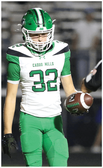 Two people in green football uniforms are on a field with one holding a football and the other preparing to receive it while players in white and black uniforms stand in the background. Descripci n generada con IA