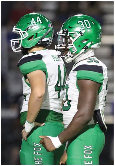 A person in a green football uniform with the number 34 is jumping over another person in a white football uniform during a nighttime game. Descripci n generada con IA