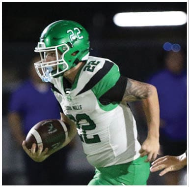 Three people in green football uniforms and helmets stand on a field at night with one person facing away from the camera and the others partially turned. Descripci n generada con IA