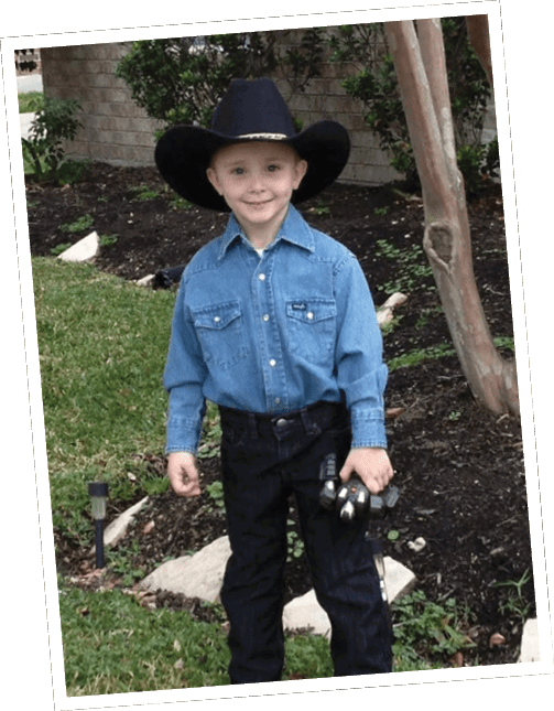 A child wearing a cowboy hat, denim shirt, and dark pants stands outdoors on grass near a garden bed holding a small toy car. Descripci n generada con IA