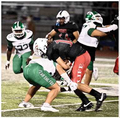 Two football players in black and red and green and white uniforms are engaged in a tackle on a field while other players watch in the background. Descripci n generada con IA