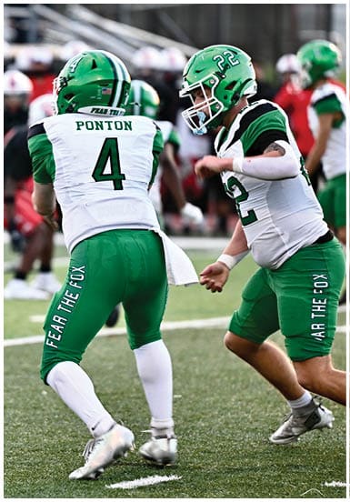 Two people wearing green helmets and white and green football uniforms are engaged in a football game on a field. Descripci n generada con IA