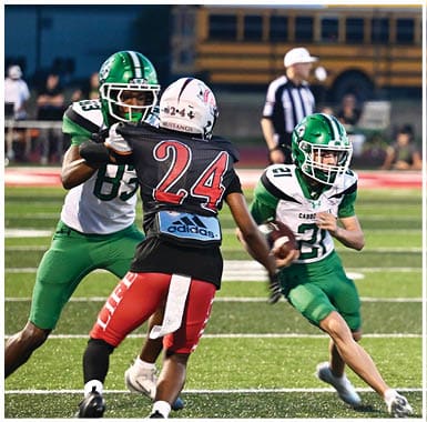 A person in a green and white football uniform is running with the ball while a person in a black and red uniform with “Mustangs" on the jersey is chasing them on a football field. Descripci n generada con IA