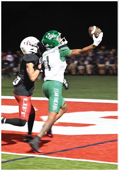 Two people in football uniforms are competing for the ball near the end zone on a field at night. Descripci n generada con IA