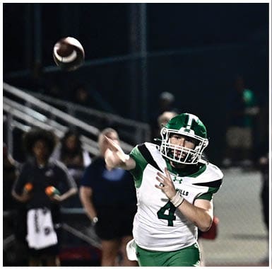 A person in a green and white football uniform is throwing a football on a field with several people standing in the background. Descripci n generada con IA