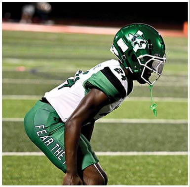 A person in a green and white football uniform and helmet is crouched in a ready position on a football field. Descripci n generada con IA