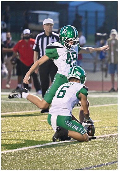 Two people in football uniforms are on a field with one holding the ball on the ground while the other prepares to kick it. Descripci n generada con IA