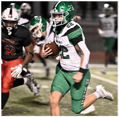 An adult wearing a green and white football uniform is running with a football while being chased by another adult in a black and red uniform on a football field. Descripci n generada con IA