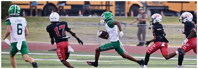 A person in green and white sportswear is running with a football while three people in black and red sportswear chase them on a football field with school buses in the background. Descripci n generada con IA