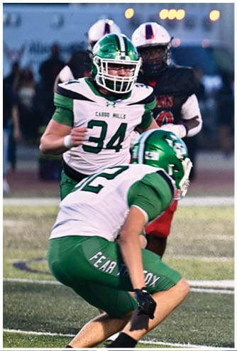 Two people in green and white football uniforms are on a field with one person in a black and red uniform in the background. Descripci n generada con IA