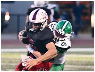 Two people in green and white football uniforms are actively playing against opponents in black and red uniforms on a field during a game. Descripci n generada con IA