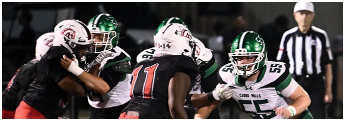People wearing football helmets and uniforms are engaged in a close physical play on a football field with a referee in the background. Descripci n generada con IA