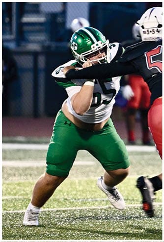 A person wearing a green and white football uniform is in a defensive stance on a football field during a game. Descripci n generada con IA