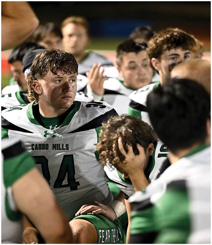 A group of people wearing football uniforms with “Caddo Mills" on the jerseys are gathered closely together, with one person in the center looking to the side. Descripci n generada con IA