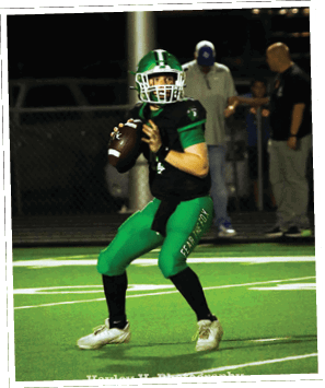 A person wearing green football pants with the words “FEAR THE FOX" printed on the side is preparing to throw a football on a turf field. Descripci n generada con IA