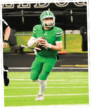 A person in a green football uniform is holding a football and appears to be preparing to throw it on a football field. Descripci n generada con IA