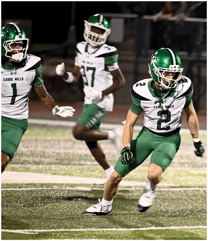 Three people in football uniforms are actively playing on a field with one person in black and red bending forward while two others in green and white approach. Descripci n generada con IA