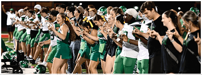 A group of people in green and white sports uniforms and cheerleading outfits are standing in a line on a field, enthusiastically clapping and cheering. Descripci n generada con IA