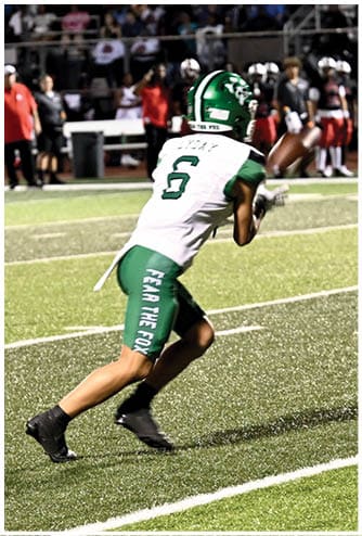 A person wearing a green and white football uniform is catching a football on a field with spectators and other players in the background. Descripci n generada con IA