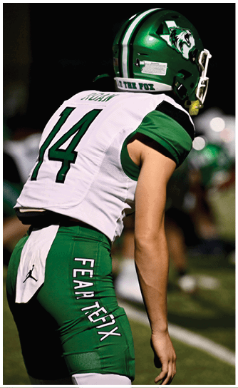 A person wearing a green and white football uniform with a helmet is standing on a field, preparing for a play. Descripci n generada con IA