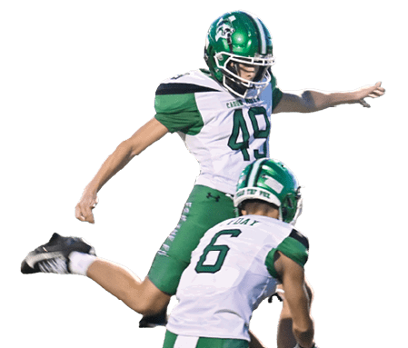 Two people in green and white football uniforms are on a field with one person holding the ball on the ground and the other preparing to kick it. Descripci n generada con IA