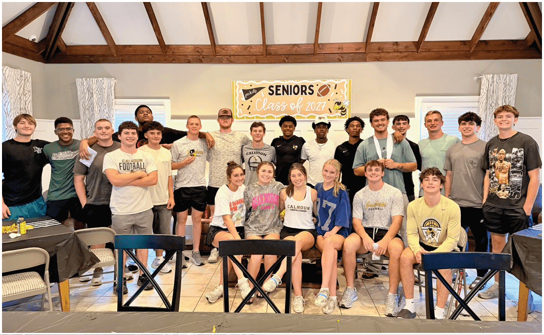 A group of people wearing casual clothing are gathered indoors in front of a banner that reads “Seniors Class of 2023" with tables and chairs in the foreground. Description generated by AI