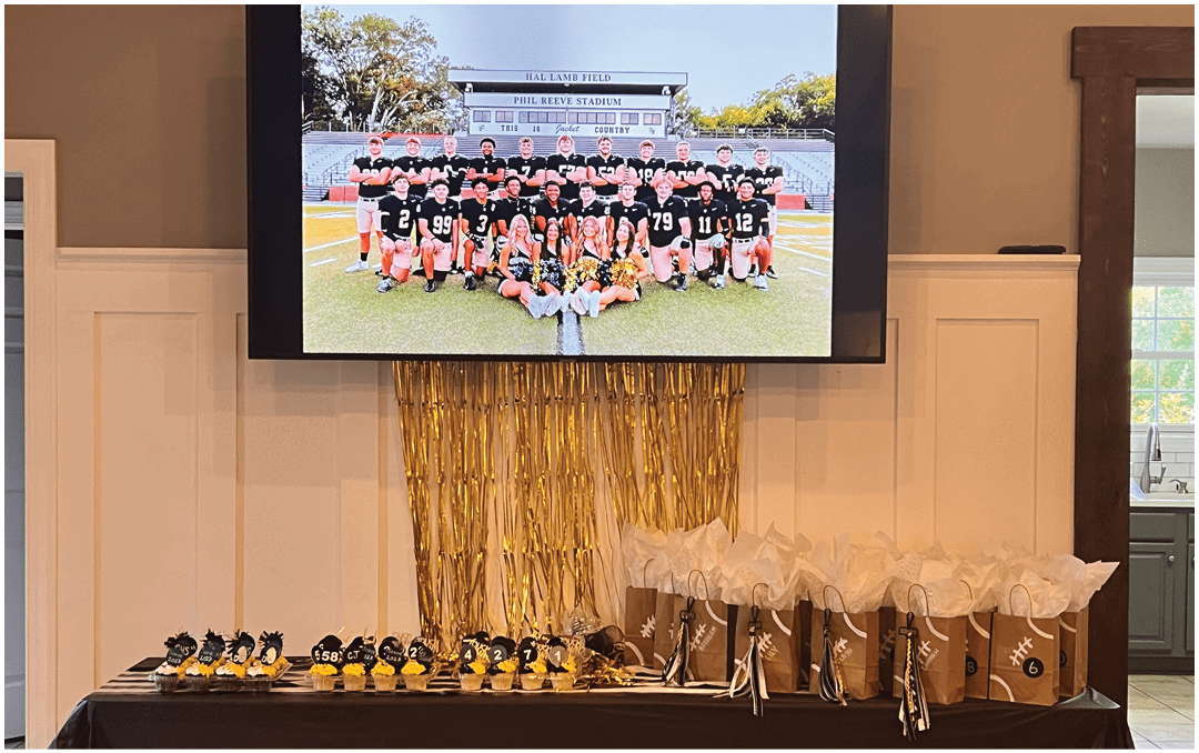 A decorated table with trophies and gift bags is set up below a large screen displaying a group photo of a football team on a field. Description generated by AI