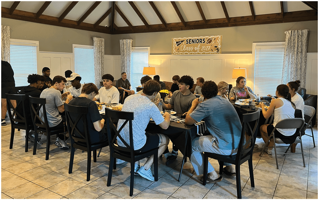A group of people sitting around a long dining table in a room with a high wooden ceiling and a banner that reads “Seniors Class of 2023" while eating and talking. Description generated by AI