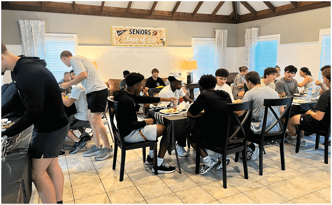 A group of people sitting and standing around tables in a room with a “Seniors Class of 2023" banner, eating and socializing. Description generated by AI