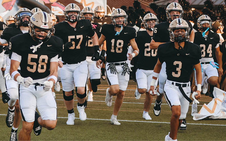 A group of football players in black and white uniforms are running onto a field through a large banner that reads “WE WILL RISE!" with a stylized "S" symbol, while a marching band stands lined up on the side. Description generated by AI