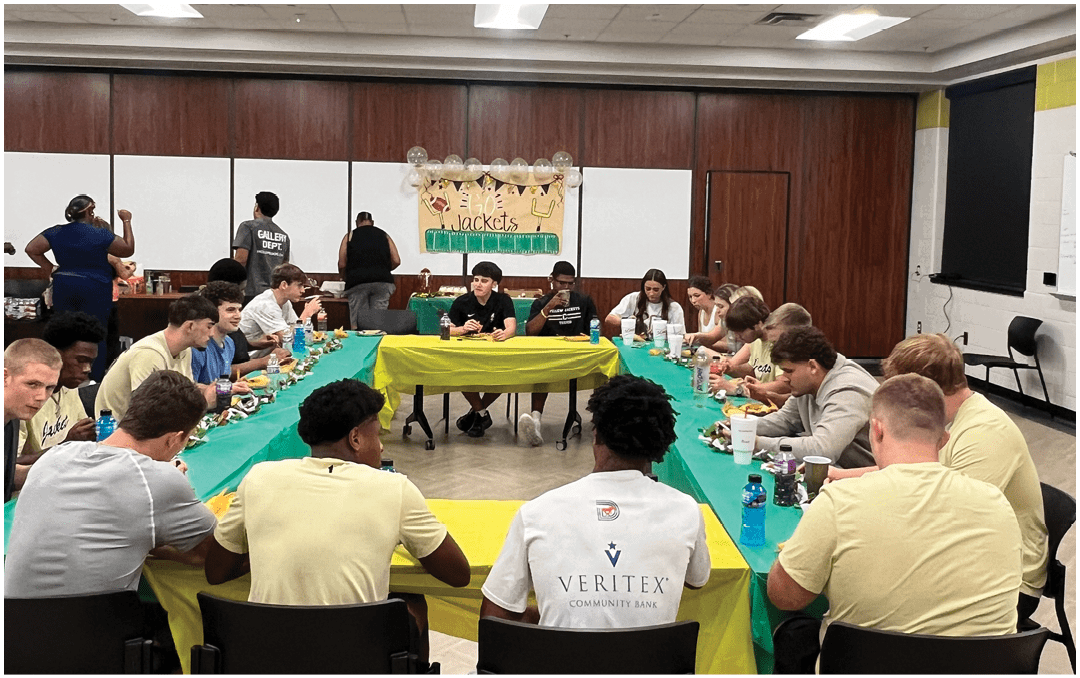 A decorated table with trophies and gift bags is set up below a large screen displaying a group photo of a football team on a field. Description generated by AI