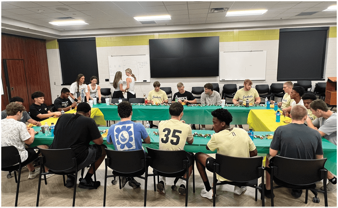 A group of people sitting and standing around tables in a room with a “Seniors Class of 2023" banner, eating and socializing. Description generated by AI