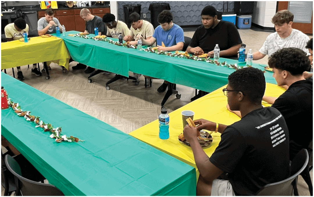 A group of people sitting around a long dining table in a room with a high wooden ceiling and a banner that reads “Seniors Class of 2023" while eating and talking. Description generated by AI