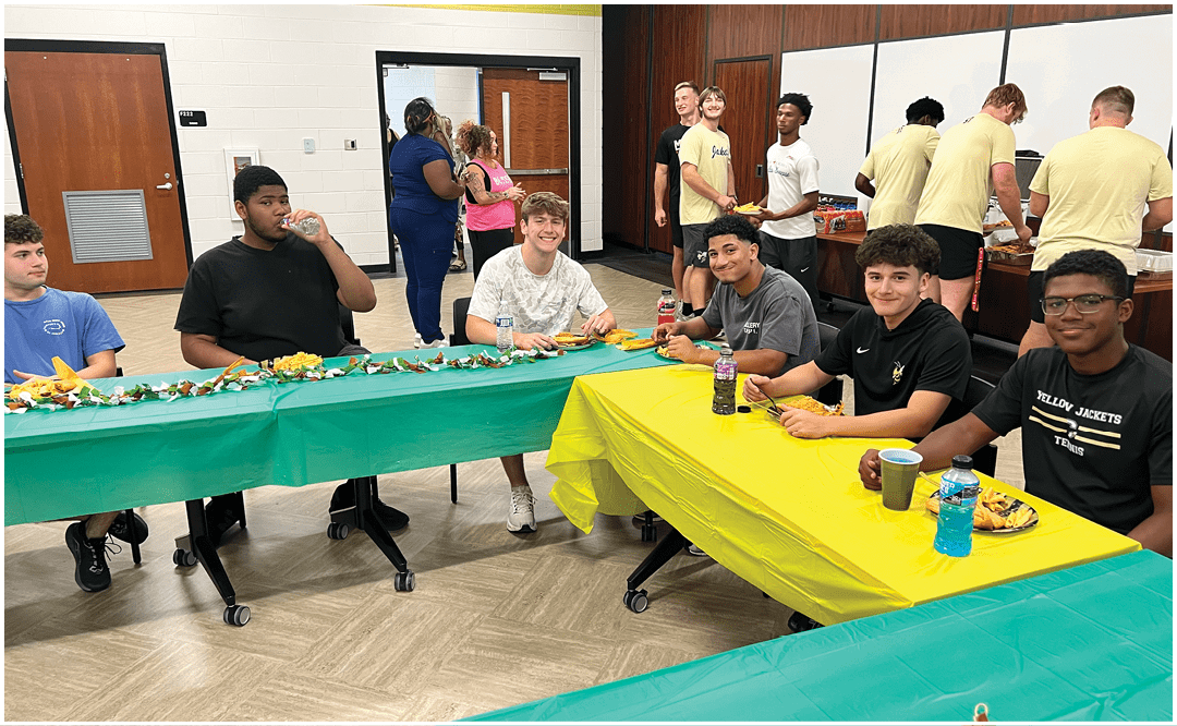 A group of people sitting and standing around tables in a room with a “Seniors Class of 2023" banner, eating and socializing. Description generated by AI