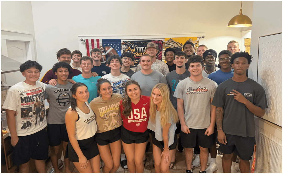A group of people wearing casual clothing are gathered indoors in front of a banner that reads “Seniors Class of 2023" with tables and chairs in the foreground. Description generated by AI