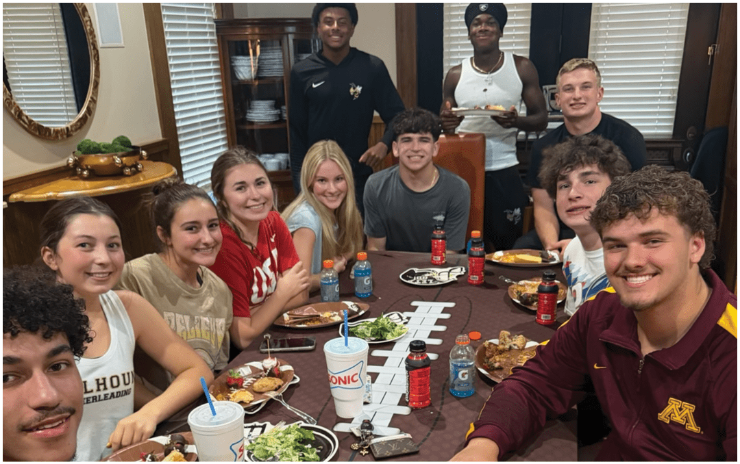 A group of people sitting around a long dining table in a room with a high wooden ceiling and a banner that reads “Seniors Class of 2023" while eating and talking. Description generated by AI