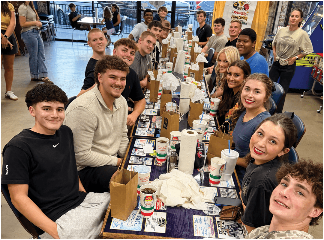 A decorated table with trophies and gift bags is set up below a large screen displaying a group photo of a football team on a field. Description generated by AI