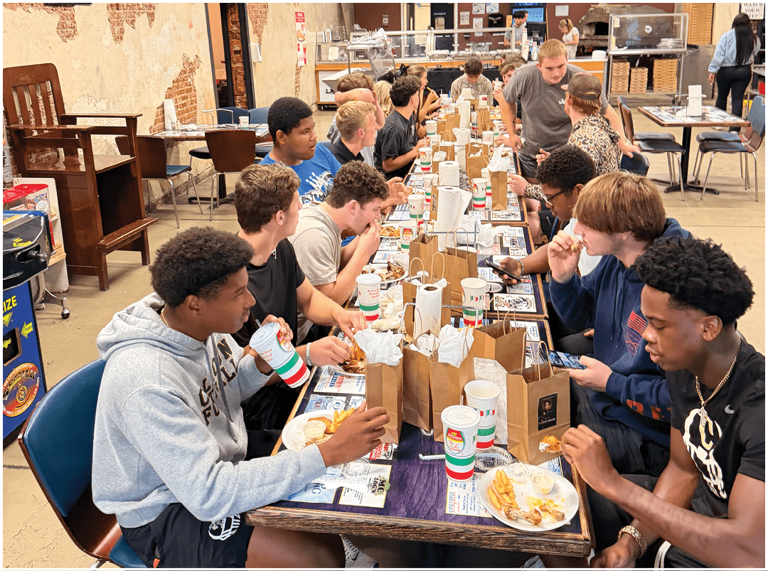 A decorated table with trophies and gift bags is set up below a large screen displaying a group photo of a football team on a field. Description generated by AI