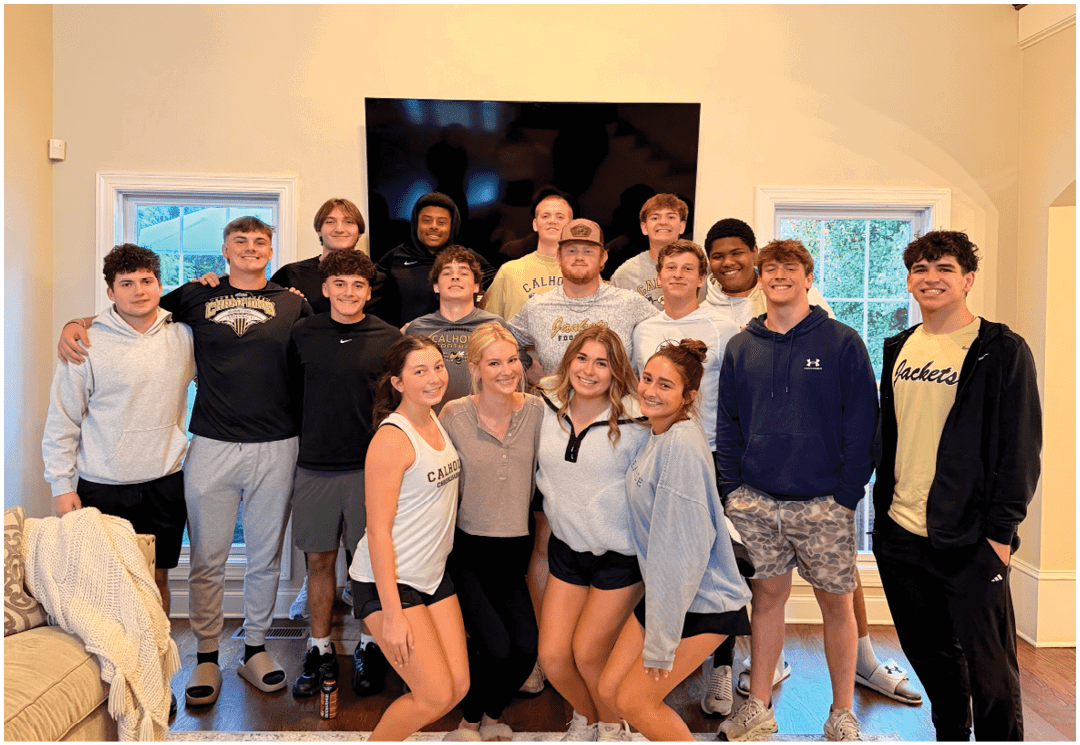 A group of people wearing casual clothing are gathered indoors in front of a banner that reads “Seniors Class of 2023" with tables and chairs in the foreground. Description generated by AI