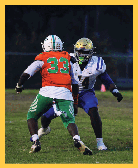 Two people in green football uniforms kneel on a field holding their helmets with heads bowed while cheerleaders stand blurred in the background. Description generated by AI