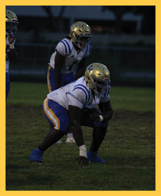Two people in green football uniforms kneel on a field holding their helmets with heads bowed while cheerleaders stand blurred in the background. Description generated by AI