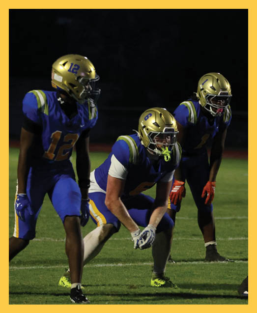 Two people in green football uniforms kneel on a field holding their helmets with heads bowed while cheerleaders stand blurred in the background. Description generated by AI