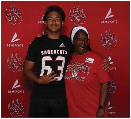 Two people stand in front of a red backdrop with sports logos wearing matching Sabercats shirts, one in black and the other in red, smiling and posing together. Description generated by AI