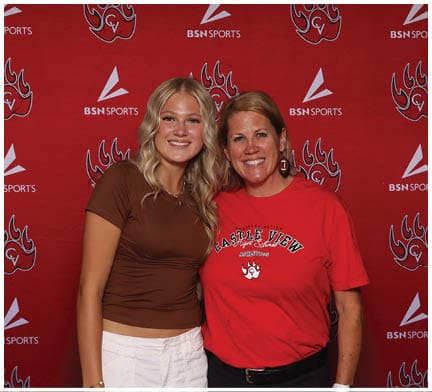 Two adults smiling and posing together in front of a red backdrop with sports logos, one wearing a brown shirt and white pants and the other wearing a red Castle View t-shirt and black pants. Description generated by AI