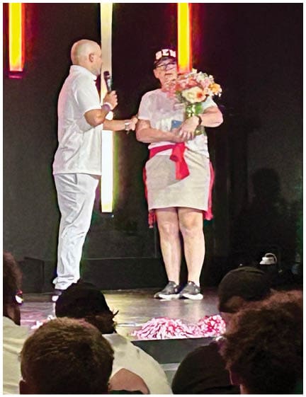 Two adults stand on a stage with one holding a microphone and the other holding a bouquet of flowers while an audience watches. Description generated by AI