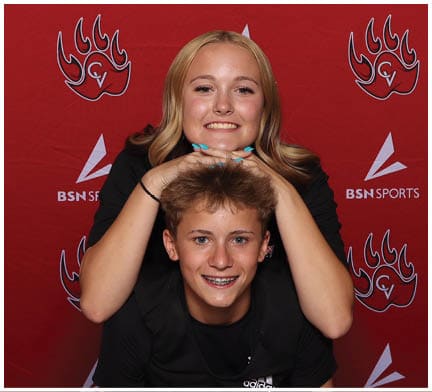 Two smiling people pose playfully with one resting their chin on the other's head against a red backdrop featuring sports logos. Description generated by AI