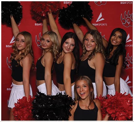 A group of smiling adults dressed in black tops and white bottoms pose cheerfully with raised pom-poms against a red backdrop with sports logos. Description generated by AI