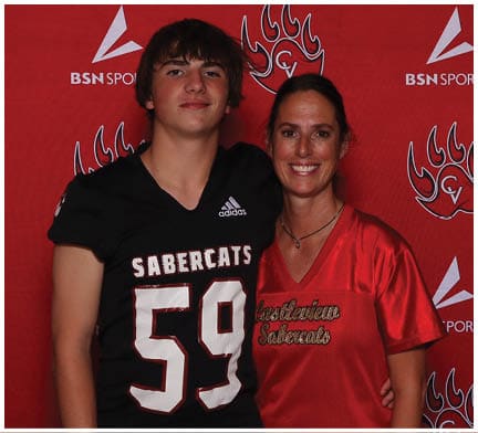 Two people stand closely together smiling in front of a red backdrop with sports logos, one wearing a black sports jersey with the number 59 and the other wearing a red sports shirt. Description generated by AI